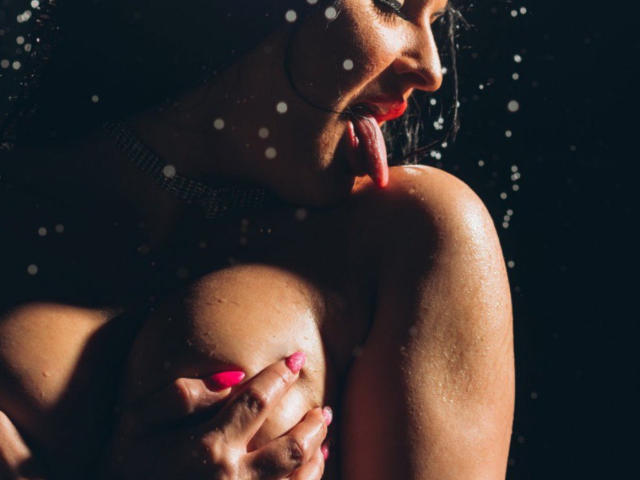 ClaireWarner's wet tits, dark skin, and pink nails are visible as she sensually licks her shoulder, with water droplets on her face and hand gripping her right boob.