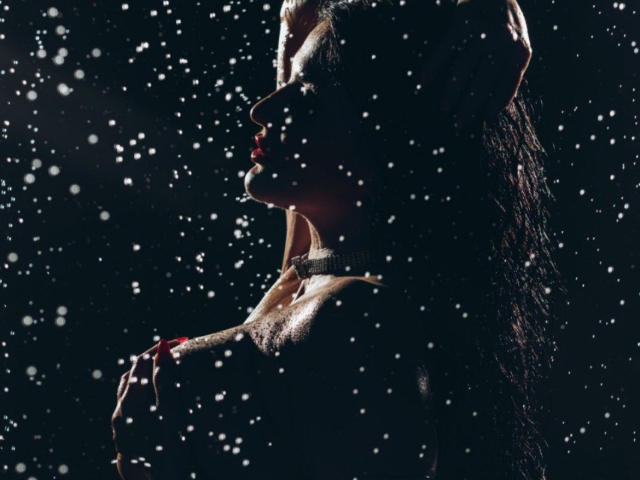 Silhouetted ClaireWarner with wet hair, dark skin, and red lipstick, holding her chest, with water droplets on her face and upper body, in a dramatic, dark setting.