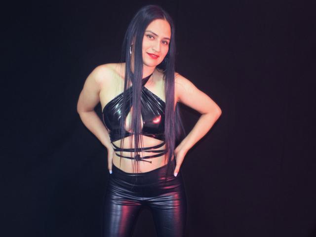 SarahMaxwell with long black hair, wearing a shiny black latex halter top and matching pants, hands on hips, smiling, showing her boobs and a slim, toned physique.