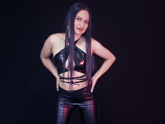 SarahMaxwell with long black hair, smiling, wearing a shiny black latex halter top and matching pants, hands on hips, standing against a dark background. Boobs, ass, tits visible.