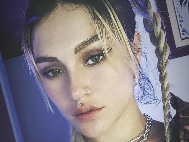Kiarrah tiene trenzas rubias, cejas pobladas, ojos verdes y un piercing en la nariz con un aro pequeño. Lleva un collar de cadena y está mirando a la cámara con un ligero puchero..