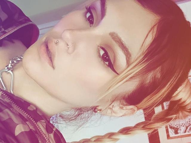 Kiarrah with red braided hair, nose piercing, and eyeliner, wearing a patterned shirt and silver chain, looking at the camera with a slight smile.