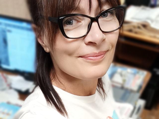 ClementineFoxy with glasses, brown hair, white shirt, smiling, small nose, light skin, subtle makeup, earrings, slightly chubby face, black-framed glasses, soft pink lips, casual setting.