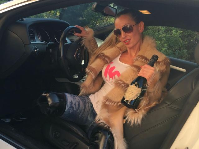 HerrinJulia-hot in car, wearing fur jacket, white tee with red design, sunglasses, holding champagne bottle, blue jeans, black boots, right hand on steering wheel, left hand on door.