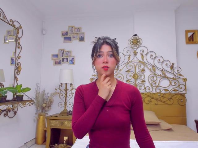 HellenBells with dark hair in a messy updo, wearing a red long-sleeve top, hand near her chin, looking surprised, standing in a bedroom with a gold ornate headboard.