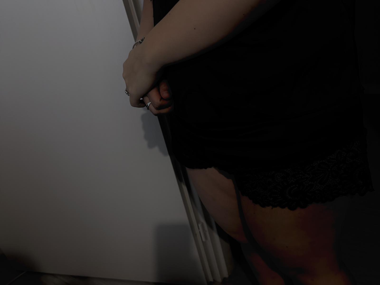 Le mani di CaramelCoupleX stringono delle mutandine in pizzo nero, rivelando un morbido ventre e un piccolo tatuaggio sul polso destro. Stanza scura e piena di ombre..