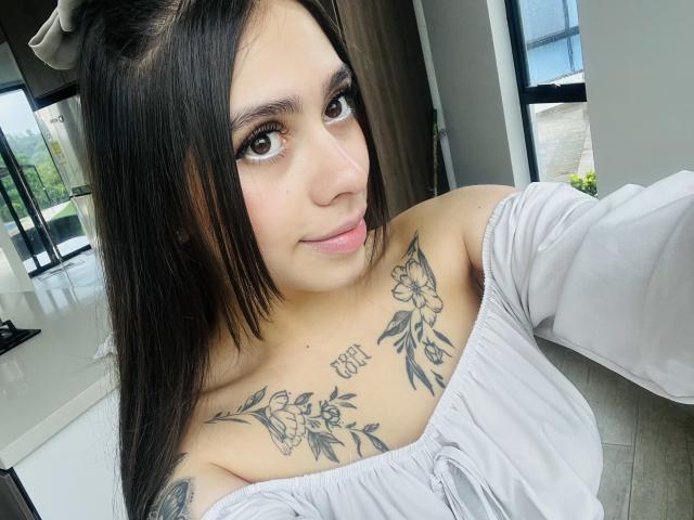 ChanelDeRosee, con cabello castaño oscuro, ojos marrones grandes y una blusa blanca con un hombro descubierto, mostrando tatuajes en su pecho y clavícula, sonriendo ligeramente, en una habitación moderna y bien iluminada..