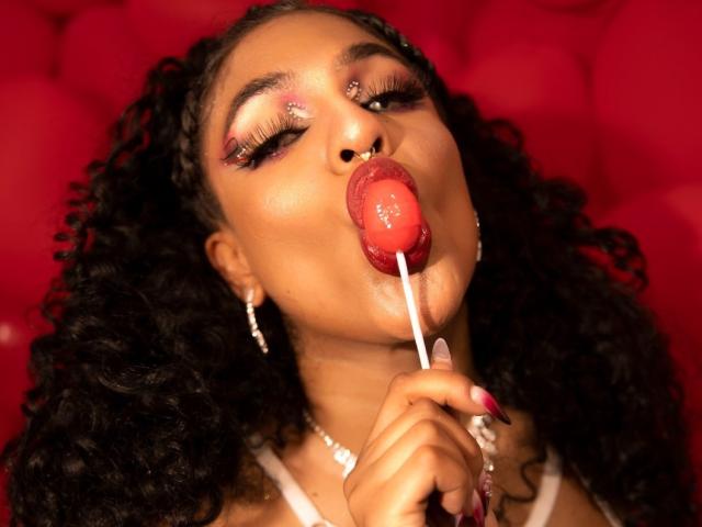 VioletHotX with curly black hair, red lipstick, and glittery eyeshadow, sucking on a red lollipop, holding it with her right hand, looking up. She's wearing white jewelry.