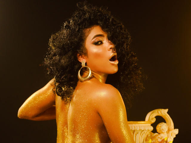 VioletHotX with curly black hair, golden glitter on her tan skin, large hoop earrings, and open mouth, holding a golden ornate vase, looking back. Boobs partially visible.