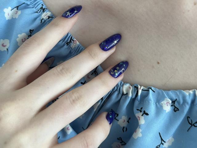 Valissia's fingers with blue, starry-nail polish on her pale skin, resting on her light blue floral dress, showing off her cute ass. Her nails are long and glossy.