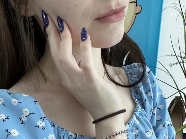 La mano di Valissia, con le unghie scure e scintillanti, tocca la sua guancia. Indossa un abito floreale blu, che mette in mostra il suo seno piccolo. Ha un braccialetto d'argento e un elastico per capelli nero..