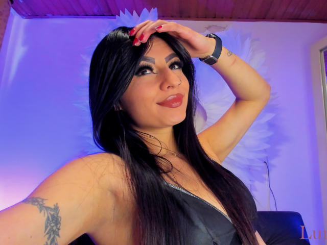 LunaCollinss with long black hair, red nails, and a tattoo on her right arm, wearing a low-cut black top, smiling, hand on head, purple-blue lighting, smartwatch on wrist.
