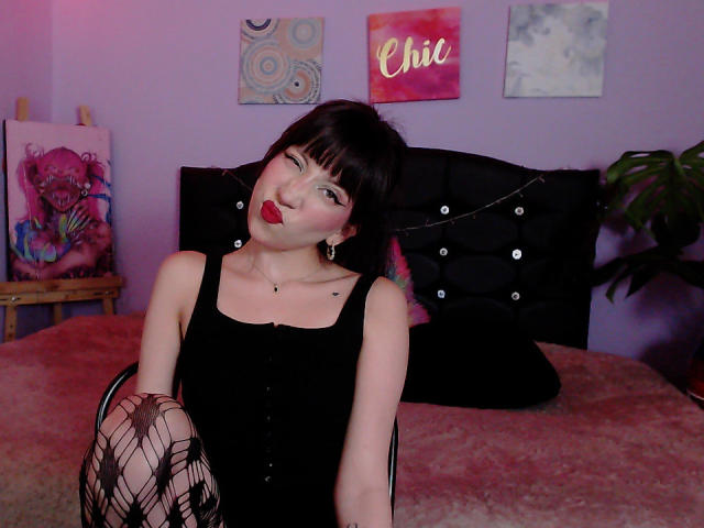 IzzysMisss with dark hair, red lipstick, black dress, fishnet knee socks, sitting on bed, winking, showing small tits, tattoo on neck, purple wall, black headboard.