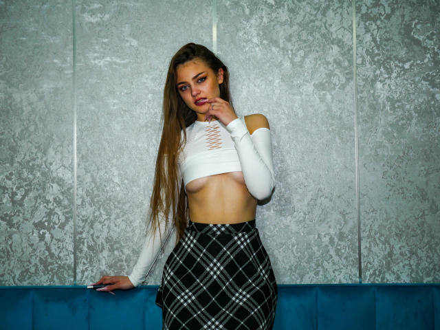 AlyssReuss with long brown hair, white crop top exposing her small tits, black and white plaid skirt, standing against a textured wall, touching her chin with a seductive look.