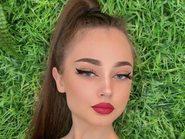AlyssReuss has red lips, dark eyebrows, long eyelashes, and a high ponytail, with green grass in the background. Her face is centered, looking slightly to the side.