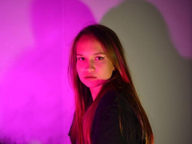 Provocateur com cabelos castanhos longos, iluminação rosa e vermelha, expressão séria, vestindo uma camisa preta, sombra em uma parede branca, luz rosa destacando o rosto, olhando por cima do ombro, maquiagem discreta..