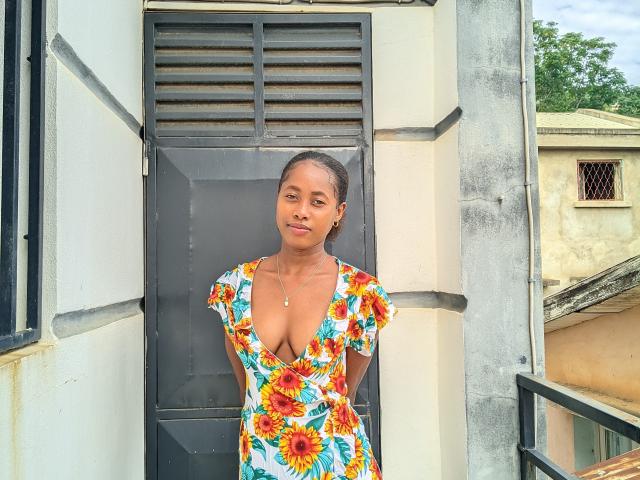 VaneOh stands in front of a door, wearing a floral dress with a deep cleavage, showing her small tits, and smiling subtly. She has a necklace and her hair's in a ponytail.
