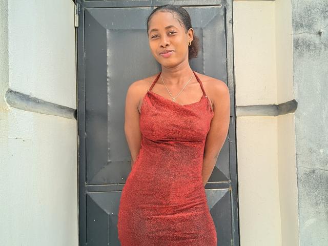 VaneOh stands in front of a door, wearing a tight red dress that highlights her ass and small tits, arms behind her back, with a slight smile on her face.