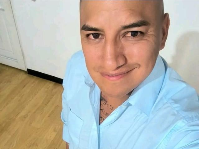 CristalDollAndJhonDS, bald, light skin, brown eyes, smiling, wearing a light blue shirt, tattoo on chest, standing on wooden floor, white wall in background.
