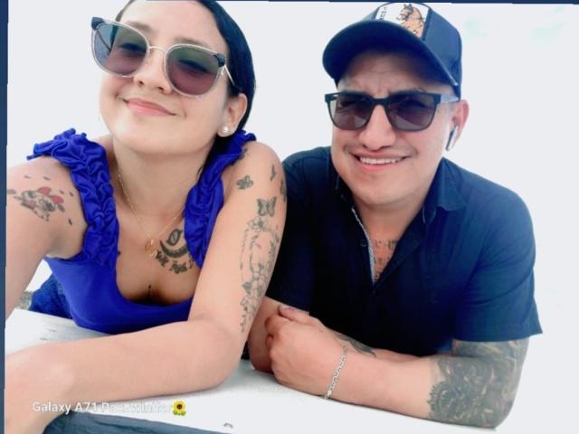 CristalDoll has tattoos, a blue ruffled top, and wears sunglasses, smiling. JhonDS in a black shirt and cap, also tattooed, with earbuds, smiles. Both are leaning on a table.