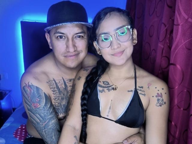 CristalDollAndJhonDS, topless with tattoos, wearing a black bikini top, standing next to JhonDS, who's shirtless, with tattoos, and a black hat, in a blue-lit room.