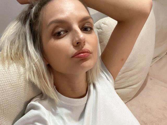 DouceAmelia with blonde hair, wearing a white tee, has her arm raised, showing a relaxed, cute face with pink lips, looking slightly to the side.