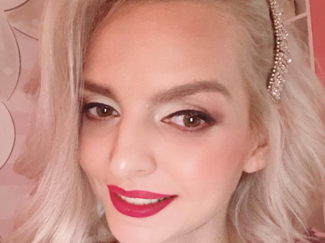DouceAmelia's face with pale skin, blonde hair, brown eyes, red lipstick, and a sparkly hairpiece. She's smiling with slightly parted lips, looking cute and sexy.
