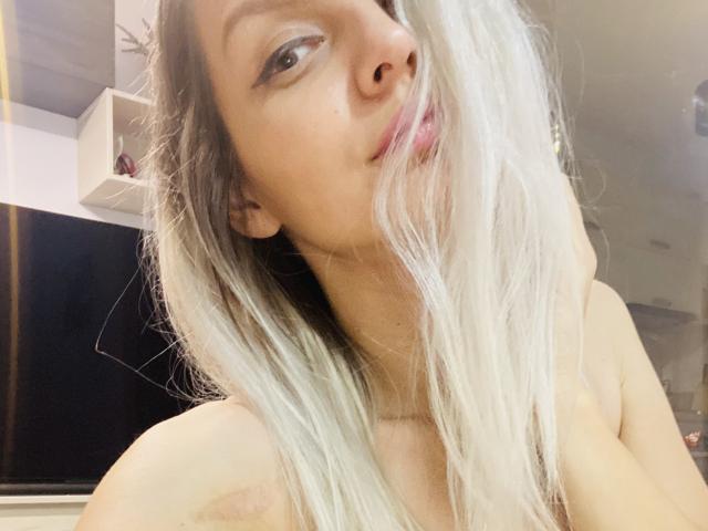 DouceAmelia with long, messy blonde hair covering her face, slightly parted pink lips, and a subtle smile, looking over her shoulder, showing bare shoulders.