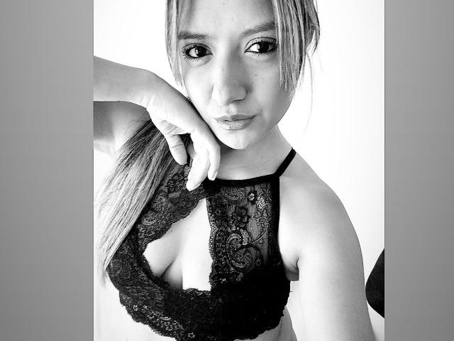 Black and white photo of ValeriaHunter69 with long hair, wearing a lace bralette, exposing her small tits, holding her hair with one hand, and giving a subtle pout.