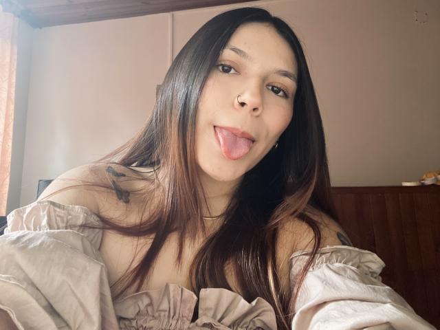 SophiaMarx with long brown hair, tongue out, wearing an off-shoulder white top, small tattoos on her left shoulder, and a nose ring, looking at the camera.