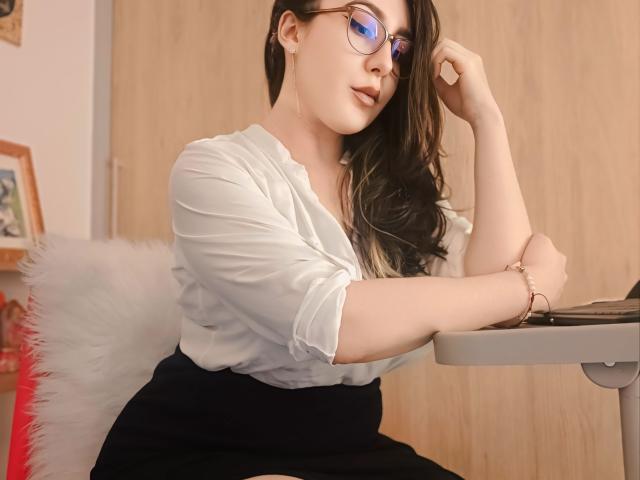 MabyLegrand with glasses, long brown hair, white blouse, black skirt, sitting at desk, left arm resting on table, right hand touching glasses, wooden door in background.