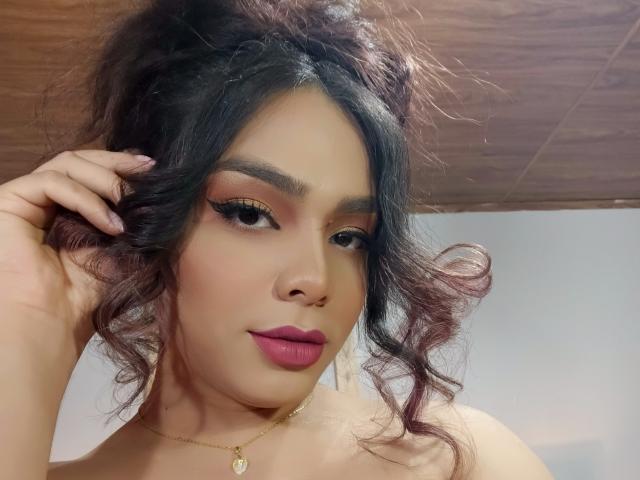LiliithBuckland has curly dark hair, bold pink lipstick, and golden eye makeup. She's touching her hair with a hand, wearing a gold necklace with a small pendant.
