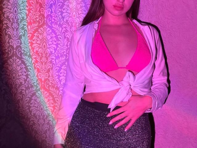 LisaAlisia con reggiseno rosa, camicia bianca annodata e gonna nera scintillante, che tiene la mano sulla pancia. I suoi seni sono visibili e si trova in un ambiente illuminato di rosa e viola..