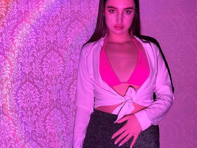 LisaAlisia com sutiã rosa, camisa branca amarrada na cintura, saia cinza com glitter, em pé, iluminação rosa-roxo, cabelo castanho longo, expressão séria, mão na saia, abdômen exposto, seios pequenos..