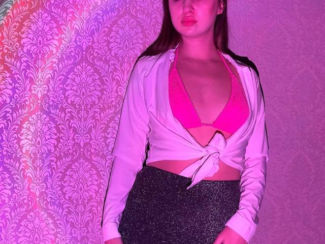 LisaAlisia con reggiseno rosa che mostra i suoi seni, giacca bianca annodata, gonna nera scintillante, illuminazione rosa e viola, labbra leggermente arricciate, capelli castani, parete bianca sullo sfondo..