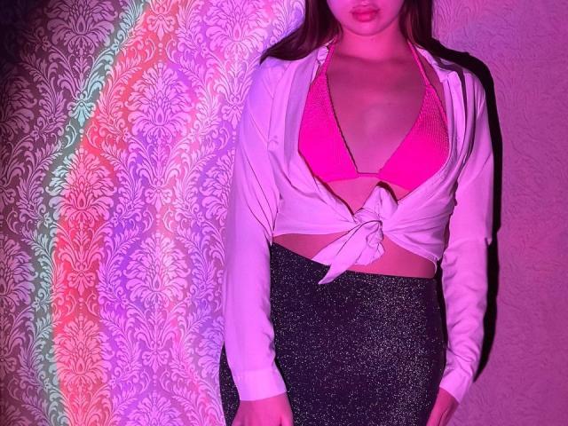 LisaAlisia with pink bra, tied white shirt, and glittery black skirt, standing, her tits visible, pink and purple lighting, her face partially in shadow, long brown hair.