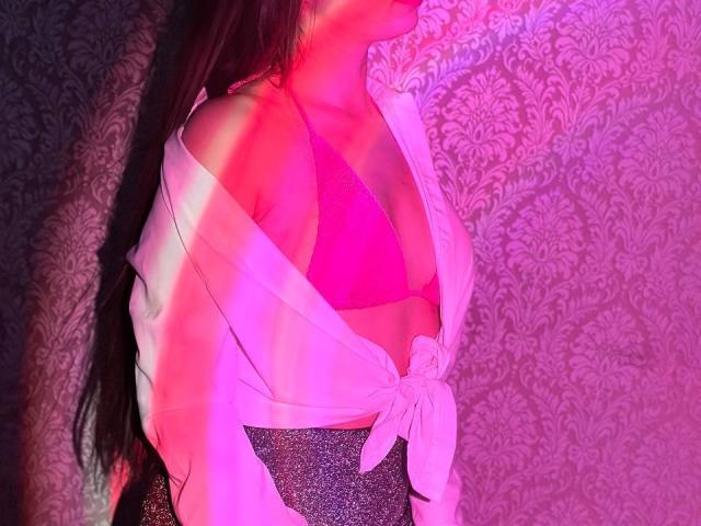 LisaAlisia, con un sujetador rosa a la vista, una camisa blanca atada debajo, una falda gris brillante, cabello negro largo, iluminación rosa y púrpura, vista parcial de lado, enfatizando sus pechos y escote..