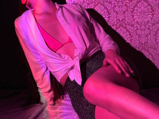 LisaAlisia con un sujetador rosa, una camisa blanca abotonada atada al frente y una falda negra con lentejuelas, sentada con las piernas cruzadas, bajo una iluminación rosa y púrpura, mostrando sus pechos y glúteos..