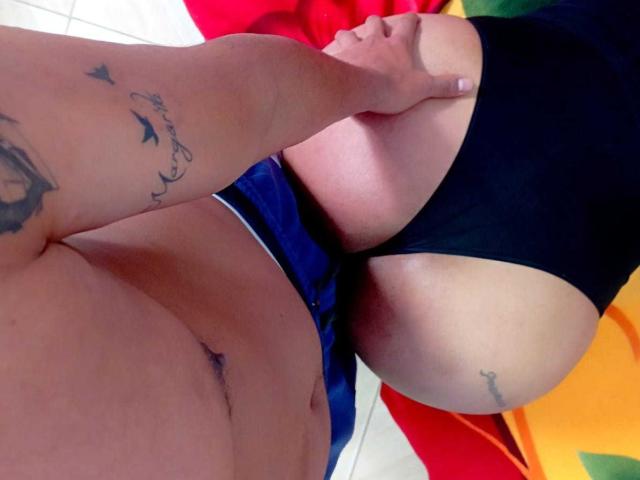 Le torse nu de LatinStyleX, orné de tatouages, tenant le postérieur de LatinStyleX, recouvert d'un string noir, montrant un tatouage sur la fesse droite, avec une main sur la fesse gauche..