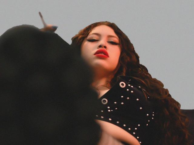 AmmyWinny with red lips, long curly brown hair, wearing a black studded top, looking down at someone with a black hairpiece in the foreground.