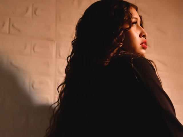 AmmyWinny, con cabello rizado oscuro, mirando hacia un lado, en una luz sombría, tiene lápiz labial rojo y lleva una blusa negra, mostrando un perfil suave y sensual..
