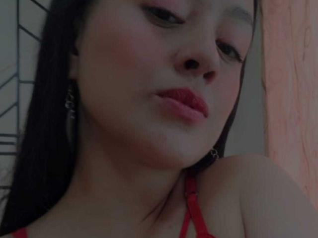 Il viso di AnnittaAngel, labbra rosse e carnose, pelle color nocciola, lunghi capelli neri, spalline rosse del reggiseno, trucco leggero, piccoli orecchini a cerchio, occhi leggermente abbassati, luce soffusa..