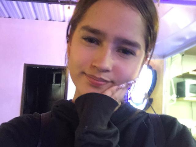 MelodyMansson has brown hair, a cute face, and is smiling with her hand on her cheek, wearing a black hoodie, in a dimly lit room.