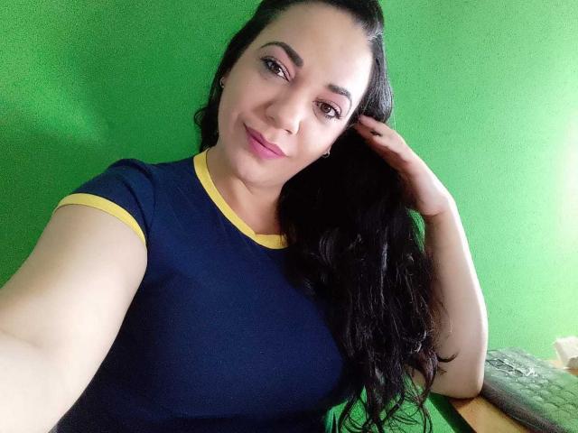 YolittS with long black hair, wearing a navy blue shirt with yellow trim, smiling, touching her hair, green background, pale skin, brown eyes, and small earrings.