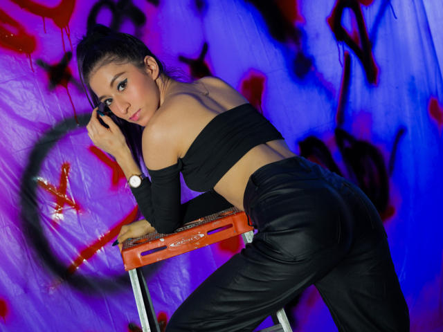 JuliaJobs with black off-shoulder top and leather pants, leaning on orange stool, looking back with a sly smile. Her ass is prominent, and she's wearing a white watch.