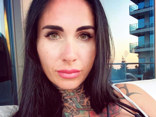 LadyPina-hot has tattoos on her neck, black hair, green eyes, and pink lipstick. Her eyebrows are dark and she's looking at the camera with a slight smile.