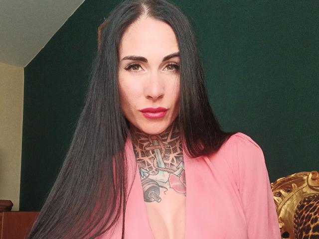 LadyPina-hot with long black hair, pink lipstick, tattooed neck, and cleavage in a pink top, looking at camera with serious expression.