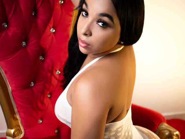EileenBenzon with dark hair, brown skin, and full lips, wearing a white lace top, sitting on a red tufted chair, looking over her shoulder with a sexy, serious expression.