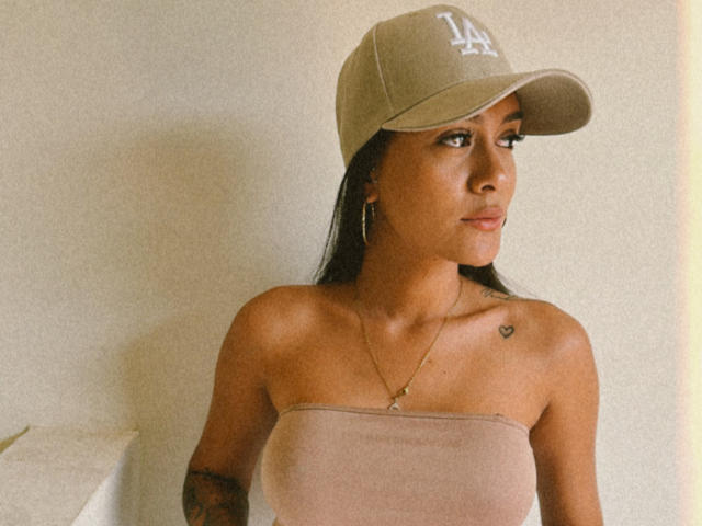 ThaliaAres with tan skin, wearing a beige baseball cap and strapless top, showing small tits and a tattoo heart on her right shoulder, looking to the side.
