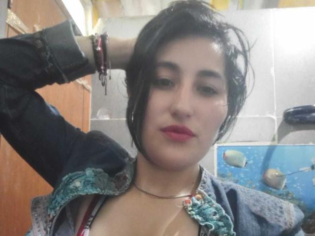 MaiGolf with short black hair, pink lipstick, wearing a blue jacket with green embroidery, hand in hair, bracelets on wrist, necklace, and visible cleavage, standing in a room.